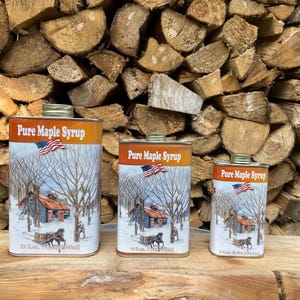 May include: Three metal cans of Pure Maple Syrup with a winter scene illustration and an American flag. The cans are different sizes: 32 fl oz, 16 fl oz, and 8 fl oz. The cans are on a wooden surface with a background of stacked firewood.