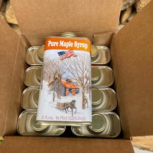 May include: A box filled with 12 cans of pure maple syrup. Each can is 8 fl. oz. and features a winter scene with a red barn, a horse-drawn sleigh, and an American flag.