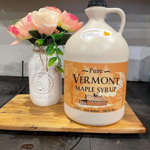 May include: A white plastic jug of pure Vermont maple syrup with a brown label. The label has a picture of a red barn and trees. The jug is sitting on a wooden cutting board. The jug is 1 gallon or 128 fluid ounces.