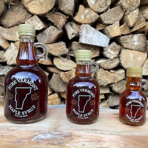 May include: Three glass bottles of maple syrup in various sizes. The bottles are clear and filled with a dark amber liquid. Each bottle has a white label with the text "Pure Vermont Maple Syrup" and an outline of the state of Vermont. The bottles are sitting on a wooden surface.
