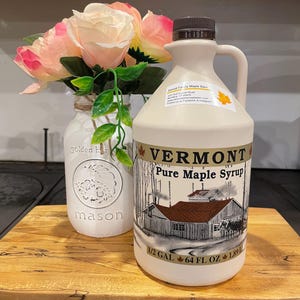 May include: A white plastic jug of Vermont Pure Maple Syrup with a red roofed house and maple trees on the label. The jug is on a wooden cutting board with a white mason jar with pink flowers in the background. The label reads "Vermont Pure Maple Syrup" and "1/2 GAL 64 FL OZ 1.89 L".