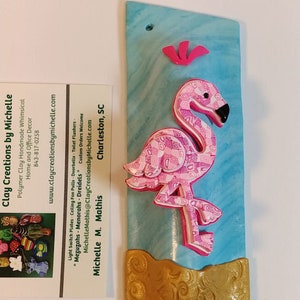 May include: A pink flamingo with a patterned design is sculpted from polymer clay and mounted on a blue and gold background. The flamingo is standing on a wave-like base. The background has a marbled effect. The piece is titled "Clay Creations by Michelle" and includes a website and phone number.