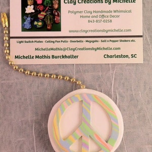 May include: A pink peace sign with a multi-colored swirl design. The peace sign is made of polymer clay and is a handmade home decor item. The business card in the image reads "Clay Creations by Michelle" and includes the website www.claycreationsbymichelle.com.