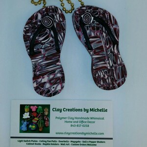 May include: A pair of flip-flop shaped polymer clay ornaments with a black and white swirl design. The ornaments are hanging from a gold chain. The business card for Clay Creations by Michelle is in the foreground. The card includes the business name, website, phone number, and a list of products.