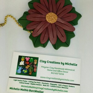 May include: A business card for Clay Creations by Michelle, a polymer clay artist. The card features a whimsical flower design and lists the artist's website and contact information. The text on the card reads: "Clay Creations by Michelle, Polymer Clay Handmade Whimsical Home and Office Decor, 843-817-0258, www.claycreationsbymichelle.com, Light Switch Plates - Ceiling Fan Pulls - Doorbells - Mezuzahs - Salt n Pepper Shakers, Cabinet Knobs - Napkin Holders - Wall Art - Custom Orders Welcome, MichelleMathis@ClayCreationsbyMichelle.com, Michelle Mathis Burckhalter, Charleston, SC."