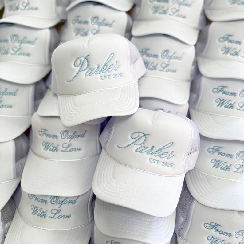 May include: A pile of white trucker hats with blue embroidered text. The text on the hats reads "Parker EST 2025" and "From Oxford With Love".