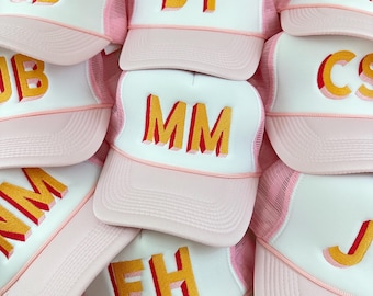 May include: A stack of pink and white trucker hats with different names embroidered on the front in red and yellow letters. The hats have a mesh back.