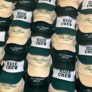 May include: A stack of green and beige trucker hats with the text "The Huies 11/16/94" and "Huie Crew" embroidered on them. The hats have a mesh back and a white front panel.
