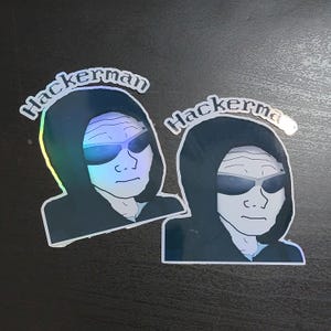 Könnte beinhalten: Zwei holografische Aufkleber mit einer Person, die einen schwarzen Kapuzenpullover und eine Sonnenbrille trägt. Der Text "Hackerman" ist auf jedem Aufkleber gedruckt.