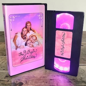 May include: A pink VHS tape case and a black VHS tape with the movie title "The Virgin Suicides". The case has a movie poster design with a group of women. The black tape is lit with a pink glow.