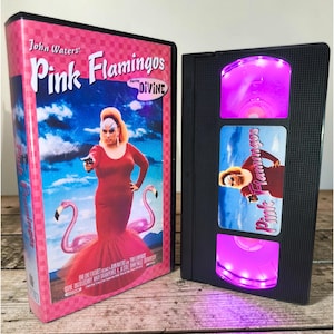Könnte beinhalten: Eine rosa VHS-Kassettenbox und ein schwarzes VHS-Kassettenlicht. Die Box zeigt den Filmtitel "Pink Flamingos" mit einer Dragqueen in einem roten Kleid und zwei rosa Flamingos. Das schwarze VHS-Kassettenlicht hat einen rosa Schein.