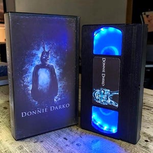 May include: A black VHS tape case and a modified VHS tape with blue LED lights. The case features the movie title "Donnie Darko" and an image of a person in a rabbit costume. The modified tape also has the movie title and a skull image.
