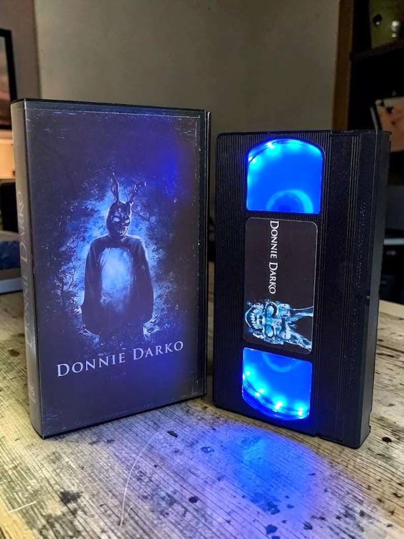 May include: A black VHS tape case and a modified VHS tape with blue LED lights. The case features the movie title "Donnie Darko" and an image of a person in a rabbit costume. The modified tape also has the movie title and a skull image.