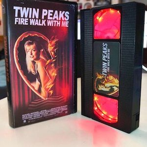 Peut inclure: Une cassette VHS noire et un boîtier noir personnalisé, tous deux avec le titre "Twin Peaks: Fire Walk With Me". La cassette VHS présente une affiche de film avec un portrait de femme. Le boîtier a des lumières LED rouges.