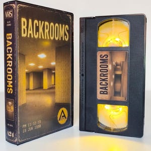 Könnte beinhalten: Ein schwarzes VHS-Band und die dazugehörige Papphülle, beide mit der Aufschrift Backrooms. Die Hülle zeigt ein gelb beleuchtetes, leeres Büroflurbild, während das Band selbst durch ein helles gelbes Licht aus dem Sichtfenster leuchtet.