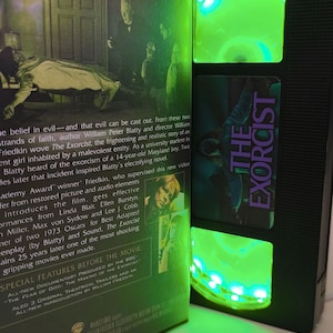 May include: A black VHS tape of "The Exorcist" movie, with a green LED light strip illuminating the tape's interior. The movie's title is in purple. The movie's cover art and text are visible.