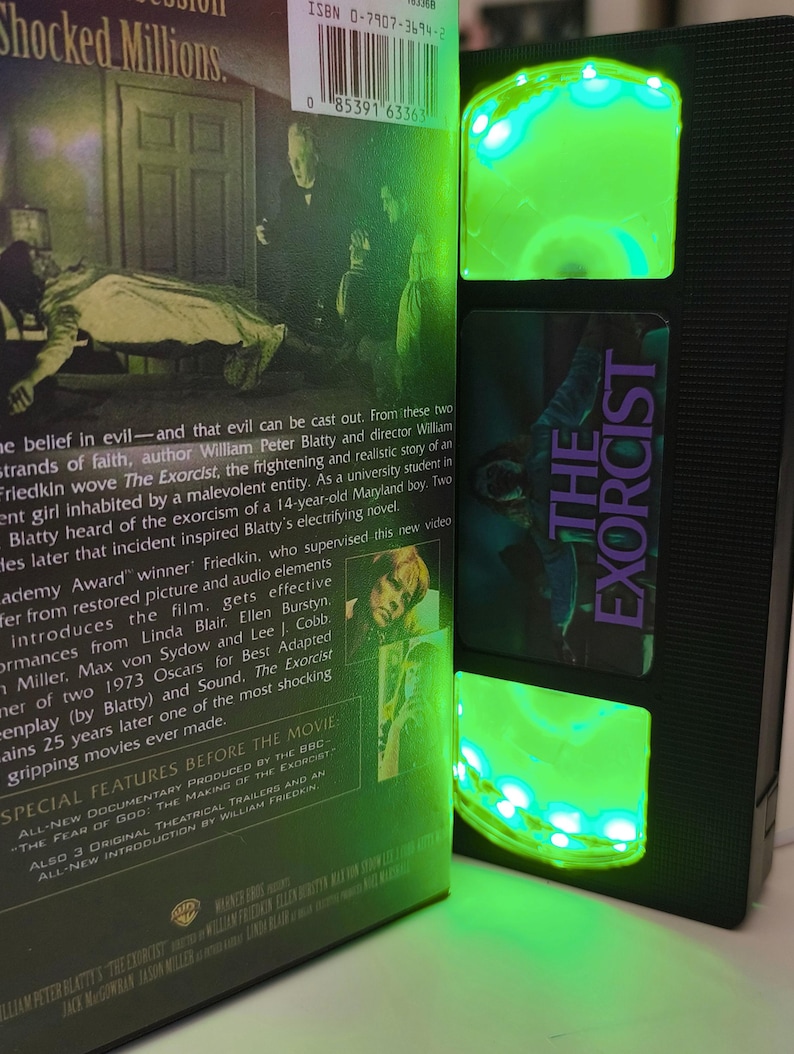 May include: A black VHS tape of "The Exorcist" movie, with a green LED light strip illuminating the tape's interior. The movie's title is in purple. The movie's cover art and text are visible.
