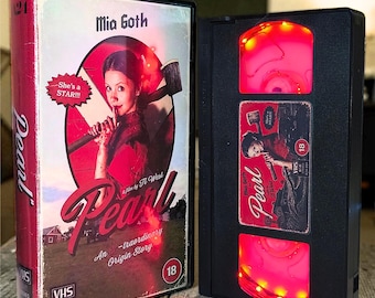 May include: Two black VHS tape cases with the movie title "Pearl" are displayed. The case on the left has a red spine and features Mia Goth on the cover. The VHS tape on the right has a red glowing light. The text "She's a STAR!!!" is visible.