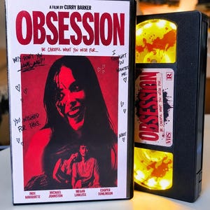 May include: A black VHS case for the film Obsession, featuring a red-toned cover with a smiling person, handwritten text, and hearts. Beside it is a black VHS tape with a yellow reel and a white label reading Obsession, Be Careful What You Wish For.