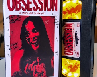May include: A black VHS case for the film Obsession, featuring a red-toned cover with a smiling person, handwritten text, and hearts. Beside it is a black VHS tape with a yellow reel and a white label reading Obsession, Be Careful What You Wish For.