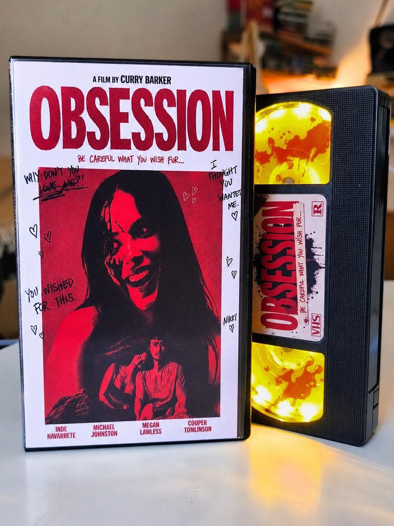 May include: A black VHS case for the film Obsession, featuring a red-toned cover with a smiling person, handwritten text, and hearts. Beside it is a black VHS tape with a yellow reel and a white label reading Obsession, Be Careful What You Wish For.