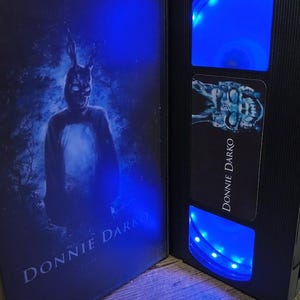 May include: A black VHS tape case and tape with the title "DONNIE DARKO" in white. The case features a figure in a rabbit mask. The tape has blue LED lights illuminating the tape window.