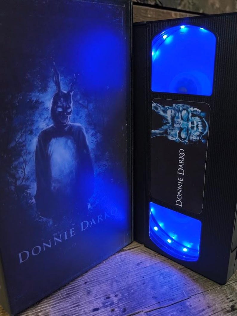 May include: A black VHS tape case and tape with the title "DONNIE DARKO" in white. The case features a figure in a rabbit mask. The tape has blue LED lights illuminating the tape window.