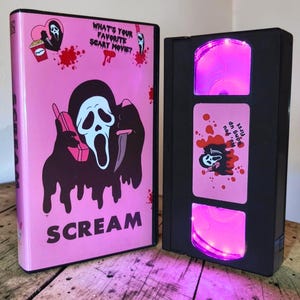 Puede incluir: Una funda de VHS rosa con el personaje Ghostface, un cubo de palomitas y el texto "WHAT'S YOUR FAVORITE SCARY MOVIE?" y "SCREAM". Al lado, una cinta VHS negra con una etiqueta rosa que dice "No, you hang up first" y luces LED de color rosa.