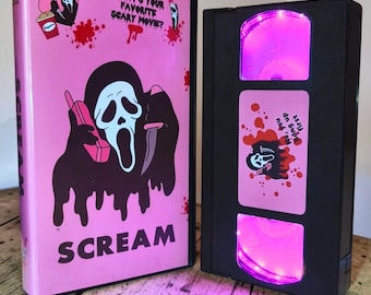 May include: A pink VHS case featuring the Ghostface character, a popcorn bucket, and the text "WHAT'S YOUR FAVORITE SCARY MOVIE?" and "SCREAM". Beside it is a black VHS tape with a pink label reading "No, you hang up first" and bright pink LED lights.