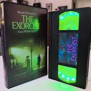 May include: A black VHS tape with the movie title "The Exorcist" and a matching DVD case. The VHS tape has a green glowing light. The DVD case features a dark green and black movie poster.