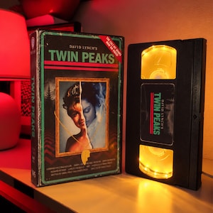 Op de afbeelding: Een Twin Peaks VHS-hoes met de naam David Lynch en een gespleten portret, naast een zwarte VHS-band die is aangepast als lamp met interne gele verlichting en een Twin Peaks-label, beide weergegeven onder warm rood omgevingslicht.