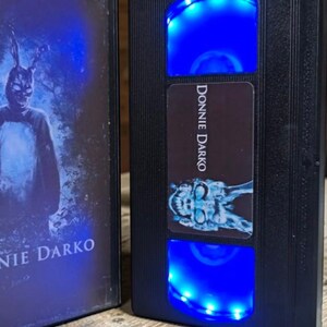 May include: Black VHS tape with the title "Donnie Darko" and a blue LED light. The cover art features a person in a rabbit costume. The VHS tape is displayed next to its case, which also features the movie title.