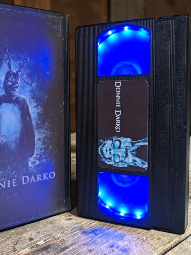May include: Black VHS tape with the title "Donnie Darko" and a blue LED light. The cover art features a person in a rabbit costume. The VHS tape is displayed next to its case, which also features the movie title.