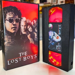 Può includere: Una videocassetta VHS del film The Lost Boys, composta da una cassetta in plastica nera con un'etichetta raffigurante un volto di vampiro e il titolo, accompagnata dalla custodia originale in cartone con sfondo rosso e il cast con occhiali.
