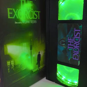 May include: A black VHS tape of "The Exorcist" with green LED lights. The movie case features a green-tinted image of a street scene with a figure. The VHS tape has a label with the movie title and a scene from the film.