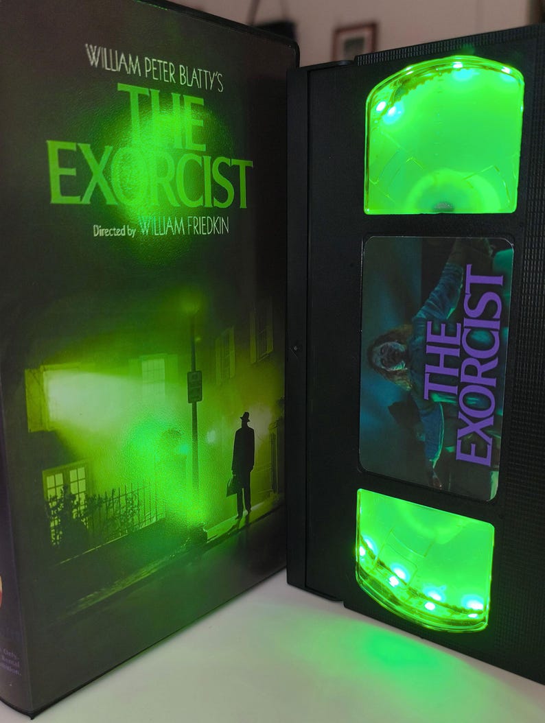 May include: A black VHS tape of "The Exorcist" with green LED lights. The movie case features a green-tinted image of a street scene with a figure. The VHS tape has a label with the movie title and a scene from the film.