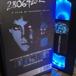 May include: A DVD case and a VHS tape featuring the movie "Donnie Darko." The DVD case has the title "28:06:42:12" and the film's title. The VHS tape is black with the movie title on the label. Both items are on a wooden surface.