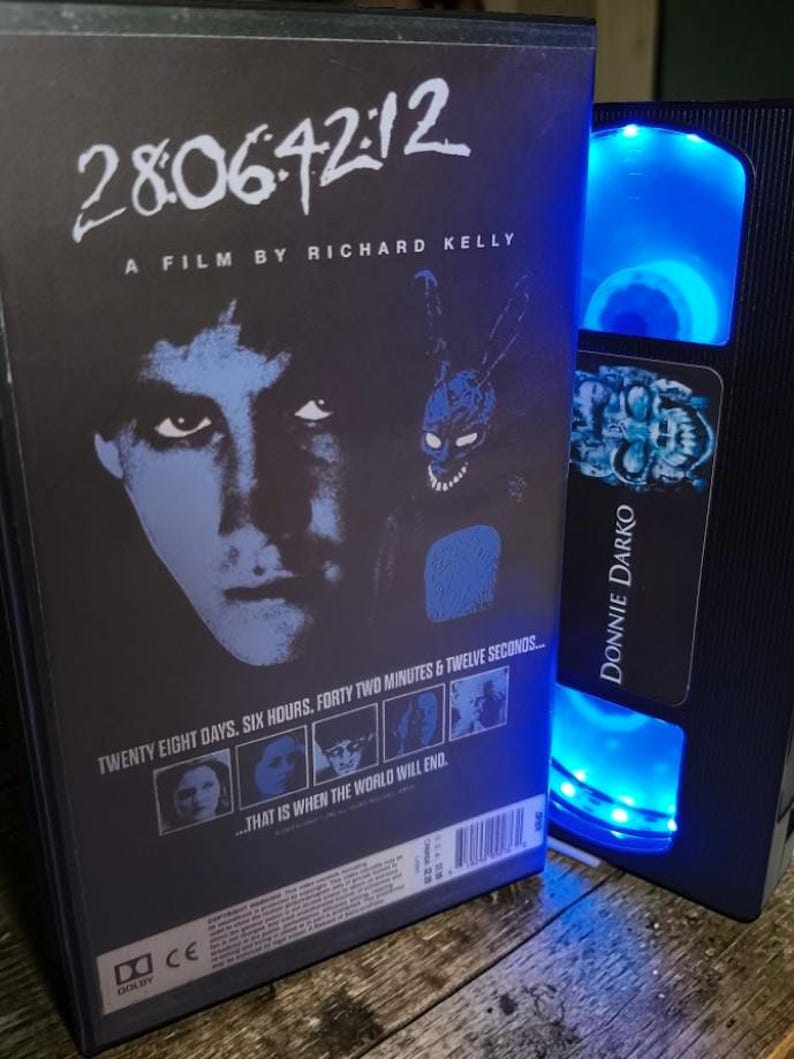 May include: A DVD case and a VHS tape featuring the movie "Donnie Darko." The DVD case has the title "28:06:42:12" and the film's title. The VHS tape is black with the movie title on the label. Both items are on a wooden surface.