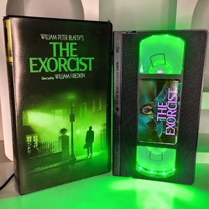 May include: A VHS tape and box set of "The Exorcist" movie. The box features the movie title in green and the silhouette of a person. The VHS tape has green LED lights.