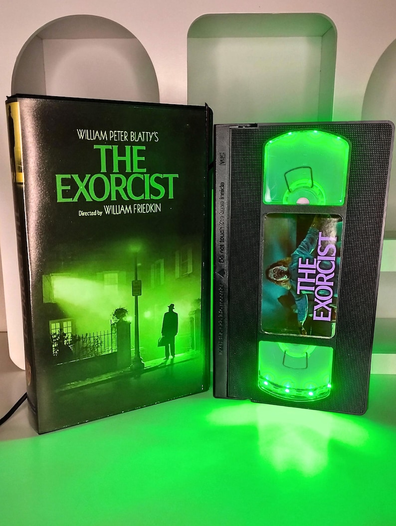 May include: A VHS tape and box set of "The Exorcist" movie. The box features the movie title in green and the silhouette of a person. The VHS tape has green LED lights.
