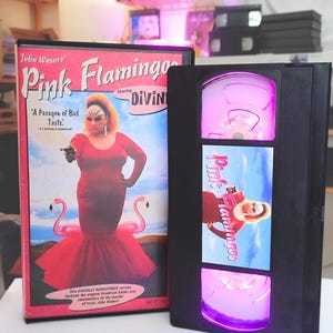 Puede incluir: Una caja y un estuche de cinta VHS negros con la película "Pink Flamingos". La carátula muestra a una persona con un vestido rojo y una pistola, con flamencos rosas. El estuche VHS tiene un interior iluminado y el título de la película es visible.