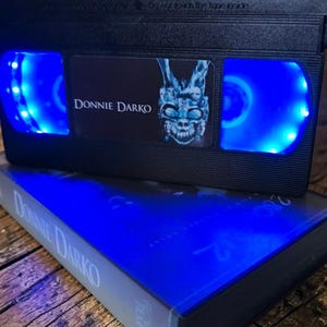 May include: A black VHS tape with the title "Donnie Darko" and a blue-lit interior. The tape is resting on top of a book with the same title. The image features a dark aesthetic with blue lighting.
