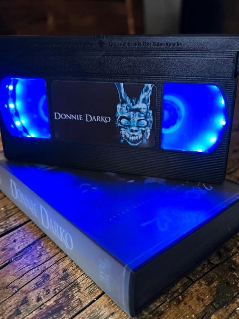 May include: A black VHS tape with the title "Donnie Darko" and a blue-lit interior. The tape is resting on top of a book with the same title. The image features a dark aesthetic with blue lighting.