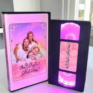 May include: A pink VHS case and a black VHS tape for The Virgin Suicides. The case features a pink-toned image of four individuals in white clothing. The tape has a custom label with the title and bright pink internal LED lighting.
