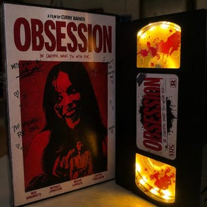 May include: A VHS tape case and a black VHS tape featuring the film title Obsession. The case cover displays a red and black graphic with the text A Film By Curry Barker and Be Careful What You Wish Fur. The tape has yellow illuminated reels.