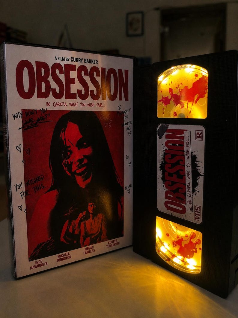 May include: A VHS tape case and a black VHS tape featuring the film title Obsession. The case cover displays a red and black graphic with the text A Film By Curry Barker and Be Careful What You Wish Fur. The tape has yellow illuminated reels.