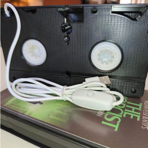 May include: A black VHS tape with a white USB cable and a switch. The tape is resting on a book with the title "The Exorcist" in green text. The tape has two white reels visible.