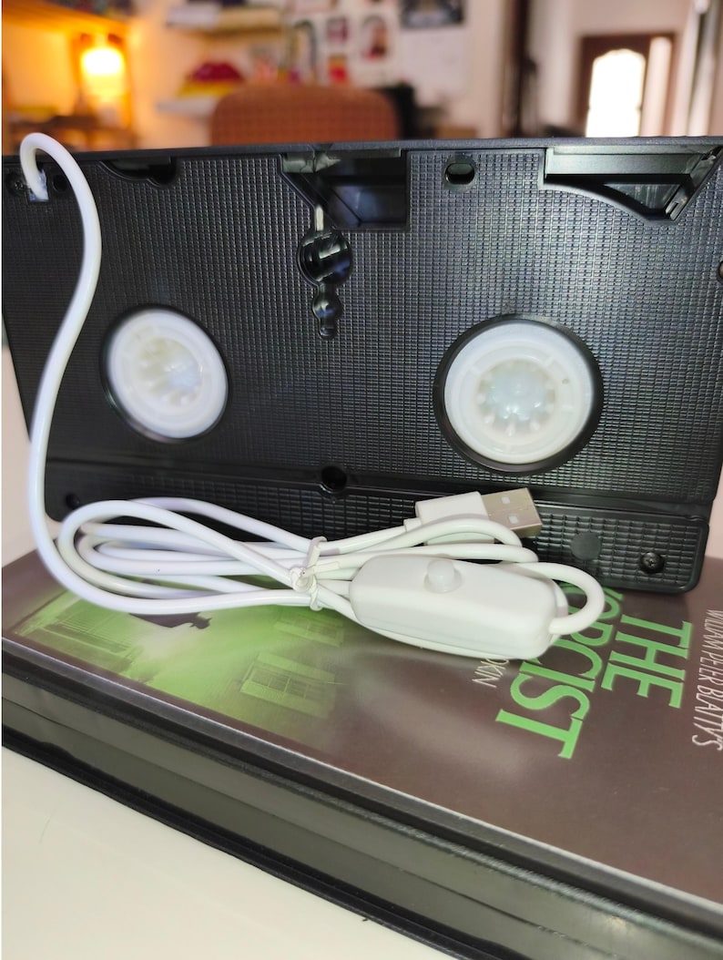 May include: A black VHS tape with a white USB cable and a switch. The tape is resting on a book with the title "The Exorcist" in green text. The tape has two white reels visible.
