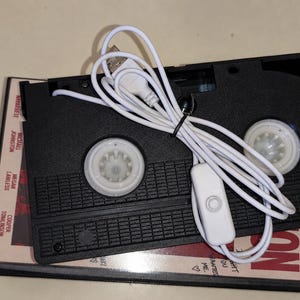 May include: A black VHS cassette tape modified with a white USB power cord and a small inline switch. The cassette rests on top of a movie case featuring the names Michael Johnston, Megan Lawless, and Cooper Tomlinson printed on the side.