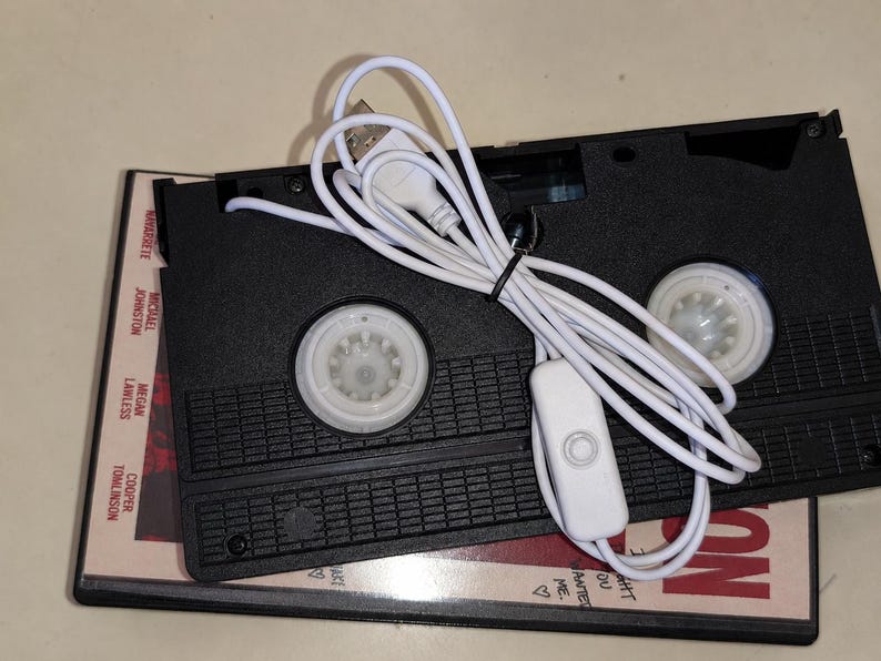 May include: A black VHS cassette tape modified with a white USB power cord and a small inline switch. The cassette rests on top of a movie case featuring the names Michael Johnston, Megan Lawless, and Cooper Tomlinson printed on the side.